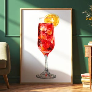 May include: Watercolor print of a cocktail in a tall glass, filled with red liquid and ice cubes, garnished with an orange slice. The artwork is framed in a light wood frame and set against a white background.