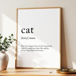 May include: A framed print with the word "cat" in a bold, black font. Below, the definition reads "[kitty] noun." The text continues with a humorous description of a cat's characteristics. The frame is a light brown color, and the print is displayed on a wooden surface.