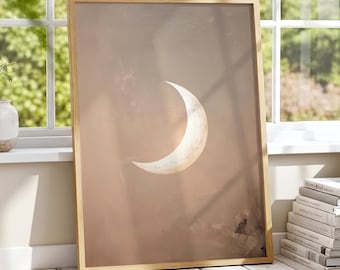 May include: A framed art print featuring a crescent moon against a muted, earthy-toned background. The artwork has a warm, neutral color palette with a gold-colored frame. The print is leaning against a window, with a stack of books visible.