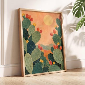 May include: A framed wall art print featuring a desert landscape with green prickly pear cacti, orange fruit, and yellow flowers under a pale yellow sun against a textured peach-coloured sky, set within a light-toned wooden frame.
