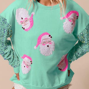 Puede incluir: Una sudadera verde menta con mangas de lentejuelas y aplicaciones de caras de Papá Noel. Las caras de Papá Noel tienen gorros rosas y barbas blancas. Las mangas están adornadas con lentejuelas turquesas y azules. Un top festivo para las fiestas.