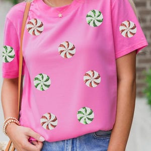 May include: A bright pink t-shirt adorned with multiple sequined peppermint candy designs in red, white, and green. The short-sleeved shirt is paired with blue denim jeans, creating a casual and festive look.