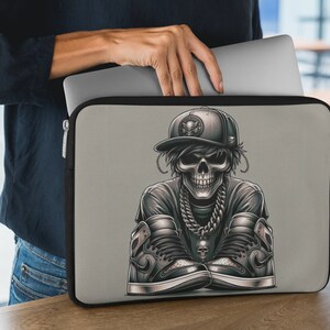 May include: A gray laptop sleeve with a black skull wearing a black and white baseball cap and a gold chain. The skull is sitting with its legs crossed and wearing black and white sneakers.