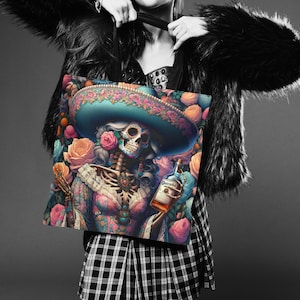May include: A tote bag featuring a colorful illustration of a skeleton woman wearing a sombrero and holding a bottle of Patron tequila. The background is filled with pink and orange roses.