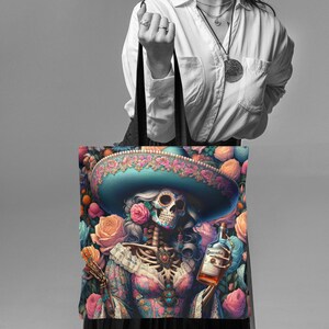 May include: A tote bag with a colorful design featuring a skeleton woman wearing a blue and pink sombrero and holding a bottle of liquor. The background is filled with flowers.