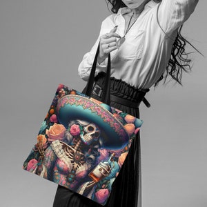 May include: A black tote bag with a colorful design featuring a skeleton woman wearing a sombrero and holding a bottle of tequila. The design includes flowers and other decorative elements.