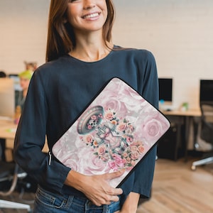 May include: A pink and white laptop sleeve with a floral design featuring a skeleton wearing a sombrero and a pink and white outfit.
