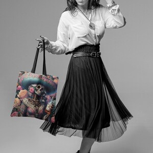 May include: A black and white photo of a person holding a tote bag with a colorful illustration of a skeleton wearing a sombrero and holding a bottle of liquor. The bag has a black strap and is made of canvas.