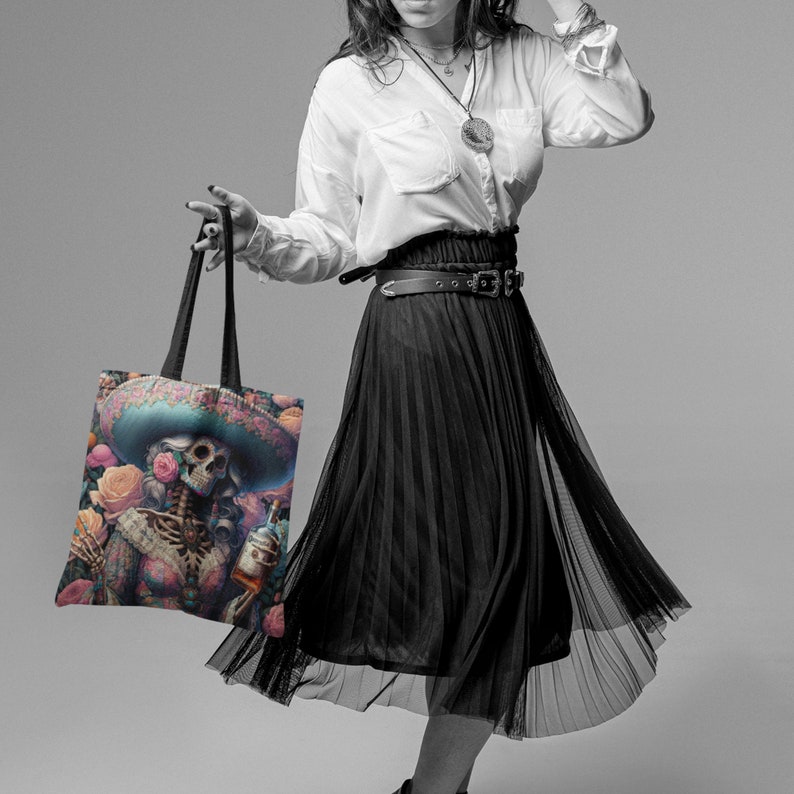 May include: A black and white photo of a person holding a tote bag with a colorful illustration of a skeleton wearing a sombrero and holding a bottle of liquor. The bag has a black strap and is made of canvas.