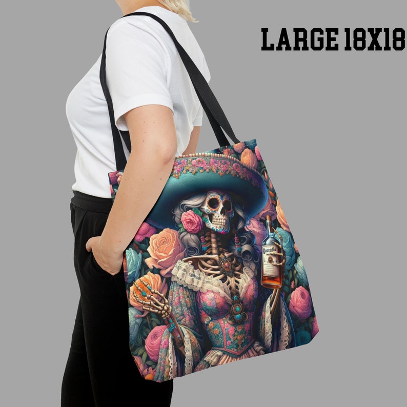 May include: A black tote bag with a colorful design featuring a skeleton wearing a sombrero and holding a bottle of liquor. The background is filled with pink, blue, and yellow flowers.