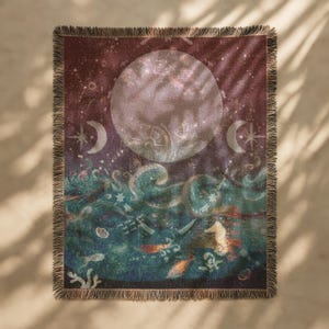 May include: A woven throw blanket with a celestial design, featuring a large moon, crescent moons, and ocean waves. The blanket has a fringed edge and a colour palette of deep red, teal, and gold. The design includes seahorses and fish.