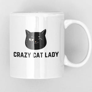 May include: A white ceramic mug featuring a black graphic of a winking cat face centred above the black text that reads CRAZY CAT LADY. The mug has a smooth finish and a curved handle on the right side, presenting a clean and simple design.