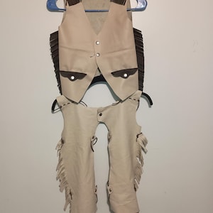 Peut inclure: Un gilet et des chaps en cuir blanc avec des franges brunes. Le gilet a une fermeture à bouton et deux poches. Les chaps ont une fermeture à sangle et sont conçus pour l'équitation.