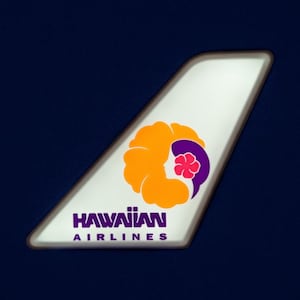 May include: A section of a white airplane tail fin with the Hawaiian Airlines logo. The logo features an orange flower design with a purple accent and a pink flower. The words "HAWAIIAN AIRLINES" are in purple text.