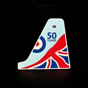 May include: A light blue sticker with a white border, featuring a design incorporating the Union Jack flag in red, white, and blue, a blue and white target, and the text "50 YEARS". The sticker has an angled shape.