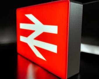 May include: A rectangular, illuminated sign featuring a bright red face with a white double-arrow symbol, commonly known as the British Rail logo. The sign has a dark, solid frame and is captured from an angled perspective.