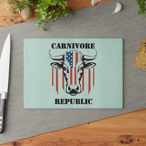 May include: A rectangular cutting board with a light blue background. The board features a graphic of a bull's head with an American flag design. The text "CARNIVORE REPUBLIC" is printed in black.