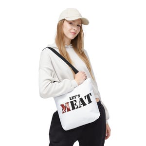 May include: White tote bag with black straps and the text "Let's Meat" in black with a red "M".