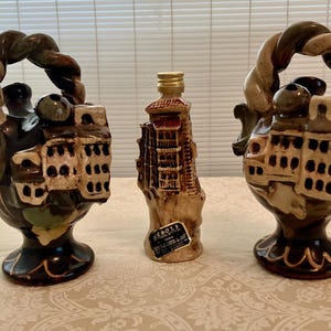 May include: Three ceramic bottle stoppers shaped like buildings with brown, green, and white glaze. The stoppers have a rope-like handle and are on a white surface.