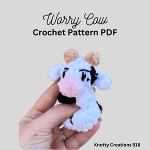 May include: A handmade white crochet cow with black spots, pink nose, and tan horns. The cow is held in a hand and the text "Worry Cow Crochet Pattern PDF" is above the cow. The text "Knotty Creations 518" is below the cow.