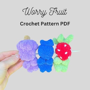Puede incluir: Tres pequeñas figuras de frutas tejidas a ganchillo en morado, azul y rojo con detalles verdes, dispuestas en un palo blanco fino. El texto superior dice Worry Fruit Crochet Pattern PDF. Las figuras tienen un aspecto texturizado, difuso y formas redondeadas.