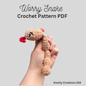 May include: A tan crochet snake with a red tongue and black and white eyes. The text "Worry Snake Crochet Pattern PDF" is displayed above the snake. The snake is held in a hand.