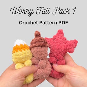 Pode incluir: Três pequenas figuras de crochê de pelúcia rotuladas como Worry Fall Pack 1 Crochet Pattern PDF. As figuras incluem um formato de doce de milho amarelo, laranja e branco, uma bolota marrom e uma folha de bordo rosa escuro, em fio macio.