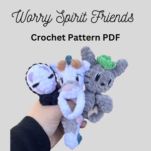 May include: Three small plush figures made of soft yarn, featuring a black and white character, a white and blue creature, and a grey figure with a green top. The text reads Worry Spirit Friends Crochet Pattern PDF in black lettering.