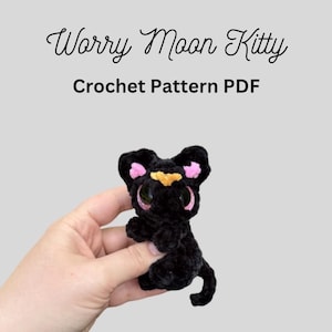 May include: A black crochet Worry Moon Kitty with pink ears and a yellow nose is held in a hand. The text "Worry Moon Kitty" and "Crochet Pattern PDF" are above the kitty.