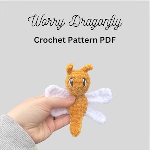 May include: A crocheted dragonfly toy in shades of orange and white. The dragonfly has large eyes and white wings. The text "Worry Dragonfly Crochet Pattern PDF" is displayed above the toy.