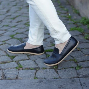 May include: Black leather slip-on shoes with a tan sole. The shoes are worn with white pants. The shoes appear to be made of a soft leather material. The shoes are a casual style.