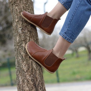 May include: Brown leather ankle boots with elastic side panels and a pull-on tab. The boots are worn by a person sitting on a tree trunk.