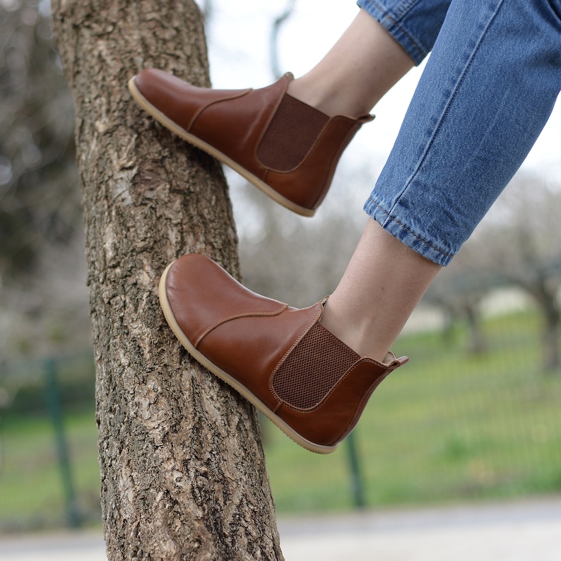May include: Brown leather ankle boots with elastic side panels and a pull-on tab. The boots are worn by a person sitting on a tree trunk.