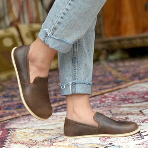 May include: Brown leather slip-on shoes with a cream-colored sole. The shoes are worn by a person wearing blue jeans.