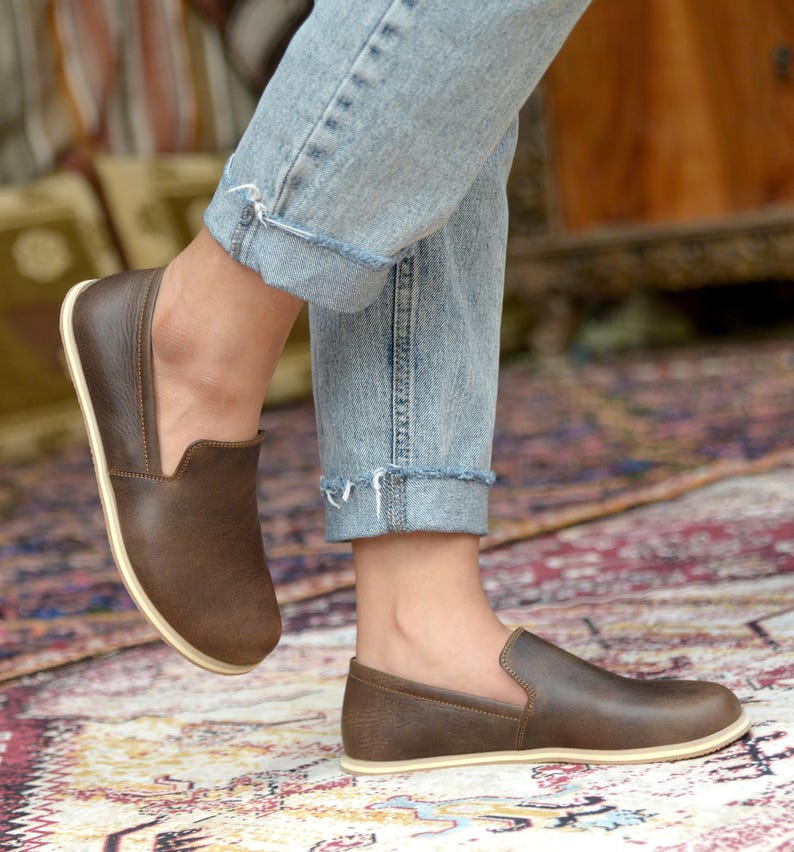 May include: Brown leather slip-on shoes with a cream-colored sole. The shoes are worn by a person wearing blue jeans.