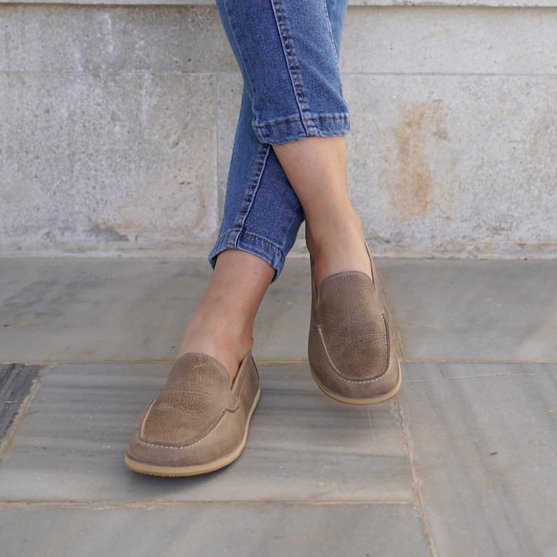 May include: A pair of light brown suede loafers with a rounded toe and a stitched design. The shoes are worn with blue jeans. The loafers have a light-colored sole.