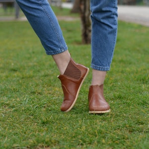 May include: A pair of brown leather ankle boots with a slight heel. The boots have a mesh panel on the back of the ankle and are worn by a person wearing blue jeans.