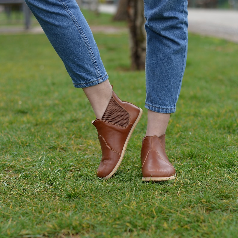 May include: A pair of brown leather ankle boots with a slight heel. The boots have a mesh panel on the back of the ankle and are worn by a person wearing blue jeans.