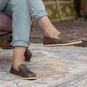 May include: A pair of brown leather slip-on shoes with a cream-colored sole. The shoes are worn by a person sitting on a chair.