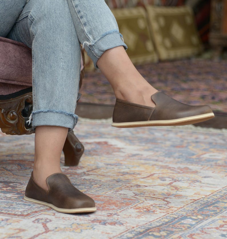 May include: A pair of brown leather slip-on shoes with a cream-colored sole. The shoes are worn by a person sitting on a chair.