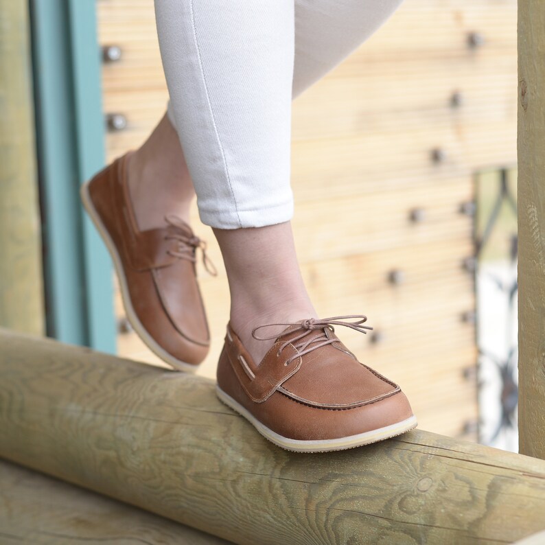 May include: A pair of brown leather boat shoes with laces. The shoes are worn by a person standing on a wooden log.