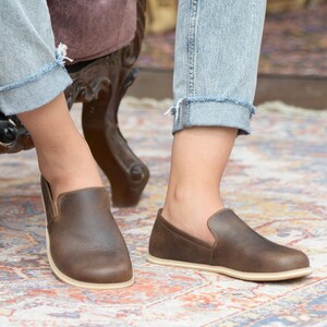 May include: A pair of brown leather slip-on shoes with a white sole. The shoes are worn by a person wearing blue jeans.