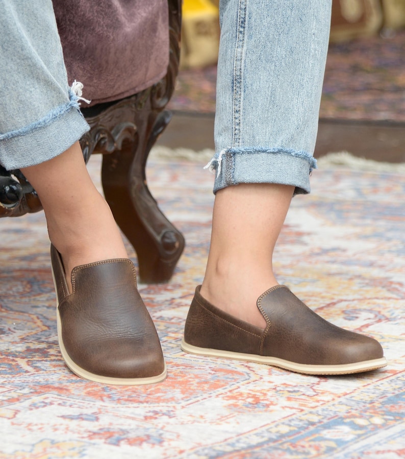 May include: A pair of brown leather slip-on shoes with a white sole. The shoes are worn by a person wearing blue jeans.