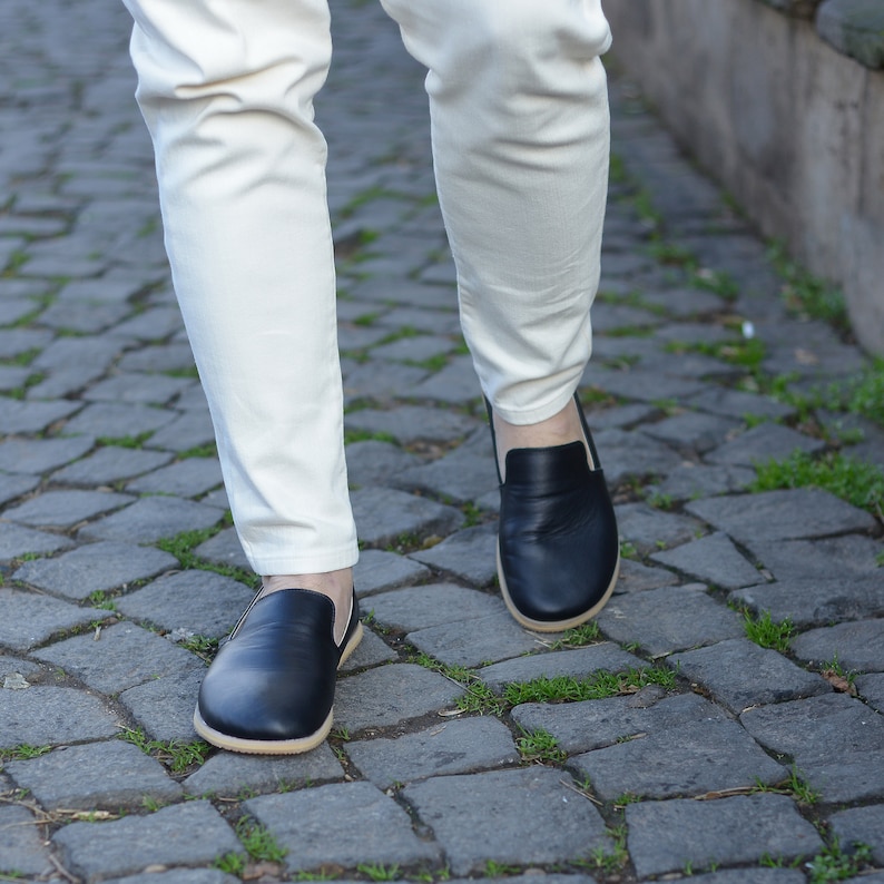 May include: Black leather slip-on shoes with a light-colored sole. The shoes are worn with off-white pants. The image is taken from a low angle, showing the shoes and the lower legs of a person walking on a cobblestone path.
