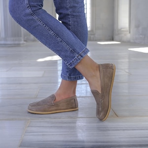 May include: A pair of taupe suede loafers with a rounded toe and a light-colored sole. The shoes are worn with blue jeans. The loafers have a classic slip-on design, suitable for casual wear.