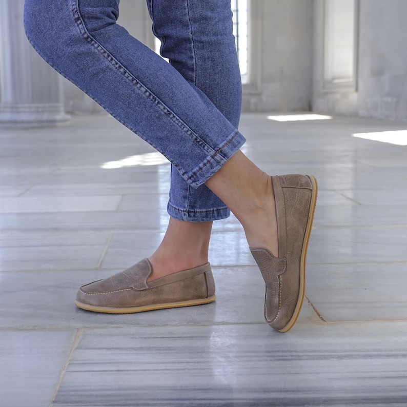 May include: A pair of taupe suede loafers with a rounded toe and a light-colored sole. The shoes are worn with blue jeans. The loafers have a classic slip-on design, suitable for casual wear.