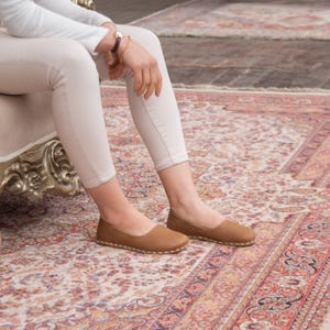 May include: A pair of brown leather flats with a stitched edge. The shoes are worn by a person sitting on a chair with white pants.
