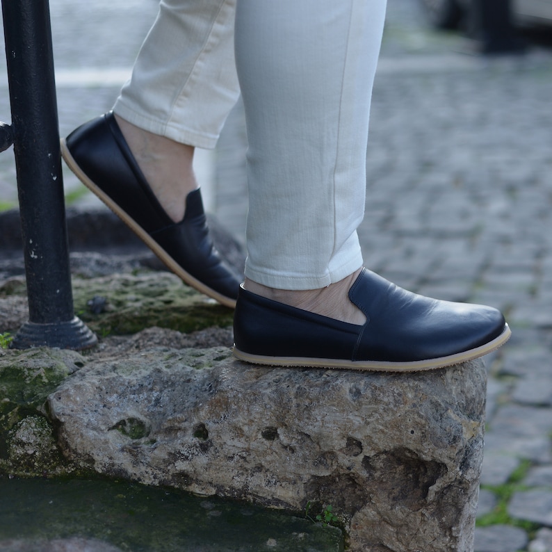 May include: Black leather slip-on shoes with a tan sole. The shoes are being worn with light-colored pants. The image is taken from a low angle, showing the shoes on a stone surface.