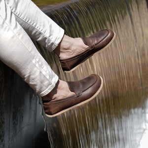 May include: A pair of brown leather loafers with a light brown sole. The loafers are worn by a person sitting on a ledge with a waterfall in the background.