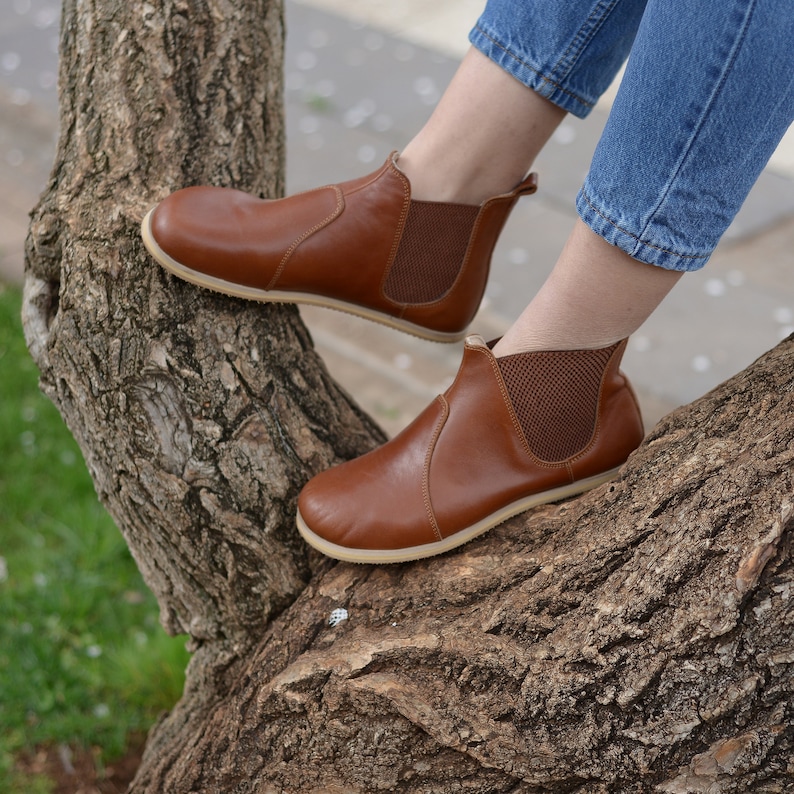 May include: Brown leather ankle boots with elastic side panels and a brown rubber sole. The boots are worn by a person sitting on a tree branch.